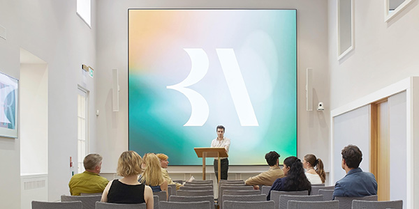 An audience sit in front of a large digital screen in a conference room